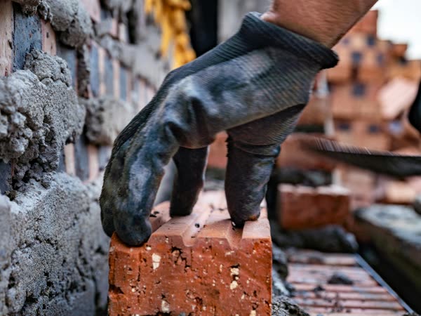 A gloved hand laying a brick on fresh mortar at a construction site